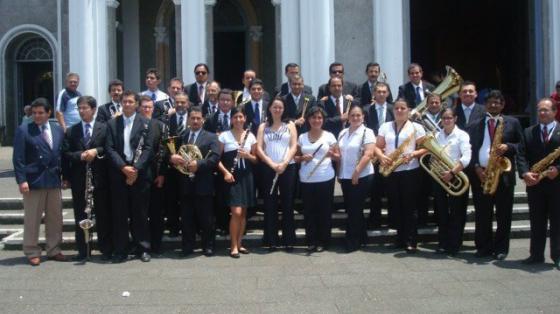 Músicos y músicas del ensamble de la Banda de Cartago frente a una iglesia grande y blanca. Todo el grupo sonríe sosteniendo sus instrumentos.