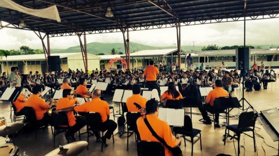 Músicos de la Banda de Conciertos de Puntarenas tocando bajo un galerón. Luz de día, público los rodea y disfruta del concierto.