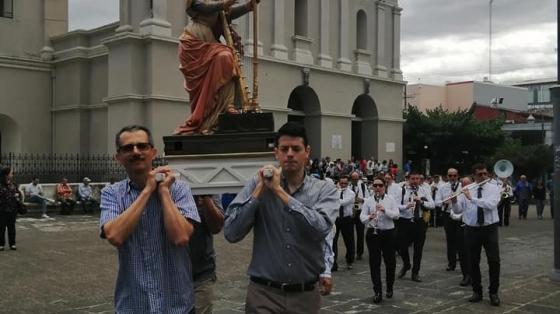 En el parque de Heredia unos hombres llevan en sus hombros un anda religiosa, atrás acompañan con música el ensamble de la Banda de Conciertos de Heredia recorriendo a pie mientras tocan para acompañar a la procesión.