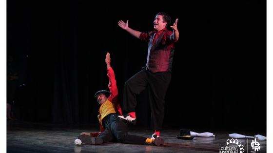 Dos chicos haciendo el acto de cierre de una presentación de circo, ambos con colores vistoso y atrás de ellos como unos pinos de boliche