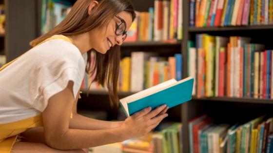 Una joven leyendo un libro, frente a unos estantes de biblioteca con libros