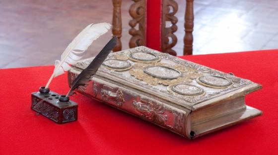 Libro antiguo cerrado junto con dos plumas para escribir, sobre un escritorio rojo