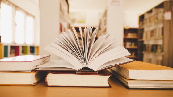 Diversos libros apilados y un libro abierto en la mesa de una biblioteca.