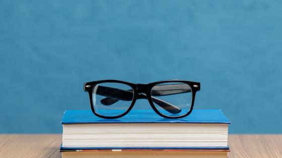 Fotografía con fondo azul, centrado se encuentran dos libros cerrados y encima un par de anteojos.