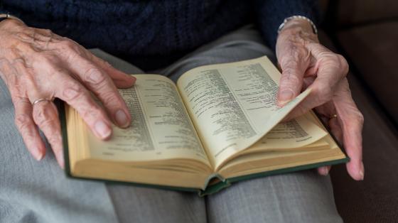 Fotografía de una persona adulta mayor leyendo un libro. En la fotografía solo se hace un plano del regazo de la persona donde se ve el libro y sus manos sosteniéndolo.  