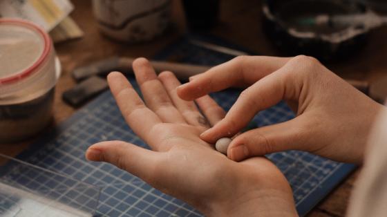 Fotografía donde se muestran las manos de una persona realizando una manualidad, en el fondo se observa un espacio con pinceles, papeles y artículos de manualidades.