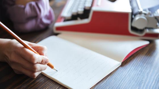 Fotografía de una persona escribiendo sobre un cuaderno abierto en blanco, al lado del cuaderno se observa una máquina de escribir de color rojo.