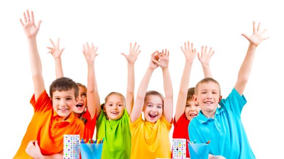 Fotografía en la que aparecen 6 niños y niñas, cada uno con una camisa de color distinto (naranja, naranja oscuro, verde, amarillo, rojo y celeste), todos los niños están sonriendo mientras levantan sus manos.