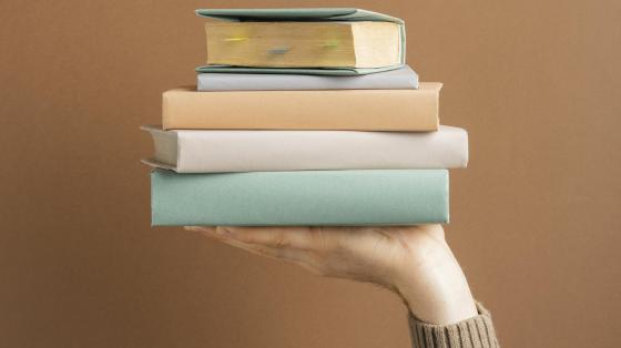 Entrega oficial Premios AICA-Costa Rica 2022 Fondo café. Una mano sosteniendo una torre de libros.