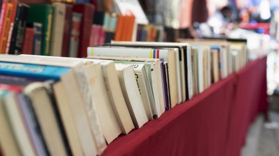 Fondo difumado. En una mesa con mantel rojo se encuentran apilados diversos libros. 