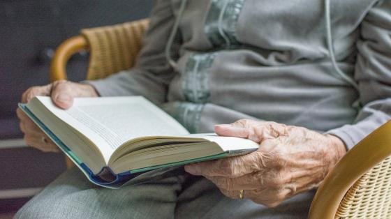 Fondo difuminado. Una persona adulta mayor se encuentra sentada sosteniendo un libro abierto.