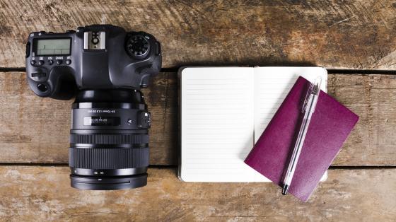 Sobre fondo de madera, a la izquierda un cámara y a la derecha un bloc de notas, un libro y un lapicero pluma. 