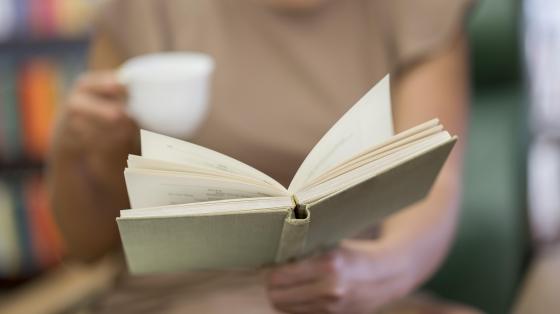 Persona de fondo difuminada con una taza y sosteniendo un libro en primer plano.