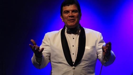 Concierto Especial "Homenaje a Ricardo Vargas" | Banda de Conciertos de Cartago Foto del Maestro Ricardo Vargas con traje blanco y fondo azul. Extiende las manos saludando.