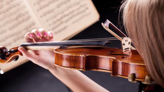 Persona sosteniendo un violón y de fondo unas partituras musicales. 