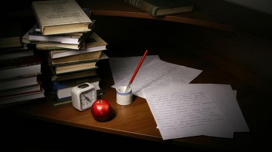 Sobre un escritorio varios libros apilados, unas hojas escritas a manos, una pluma para escribir, un reloj y una manzana