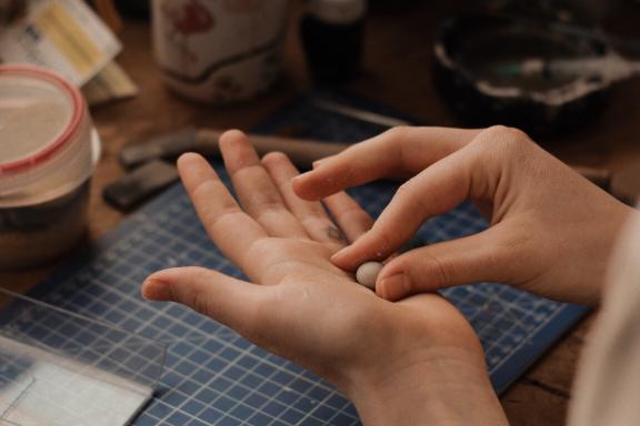 Fotografía donde se muestran las manos de una persona realizando una manualidad, en el fondo se observa un espacio con pinceles, papeles y artículos de manualidades.