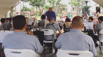 Músicos de la Banda de Conciertos de Alajuela sentados, tocando un concierto en el kiosco del parque central de Alajuela, dirigidos por Do Gabriel Campos. Concierto de día en el que se aprecia al público disfrutando del concierto.