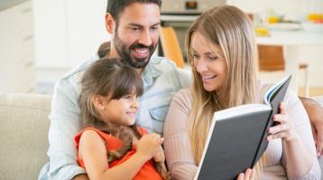 Padres alegres con una niña, sentados en el sofá de la casa, leyendo un libro y sonriendo