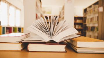 Diversos libros apilados y un libro abierto en la mesa de una biblioteca.