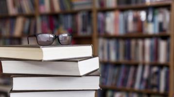 Fotografía con un fondo en segundo plano de estaterías de libros. En el frente de la fotografía se encuentran 4 libros apilados y encima de estos un par de gafas.