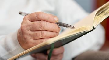 Fotografía de una toma cercana a las manos de una persona adulta mayor, sosteniendo un lápiz y cuaderno, tomando notas.